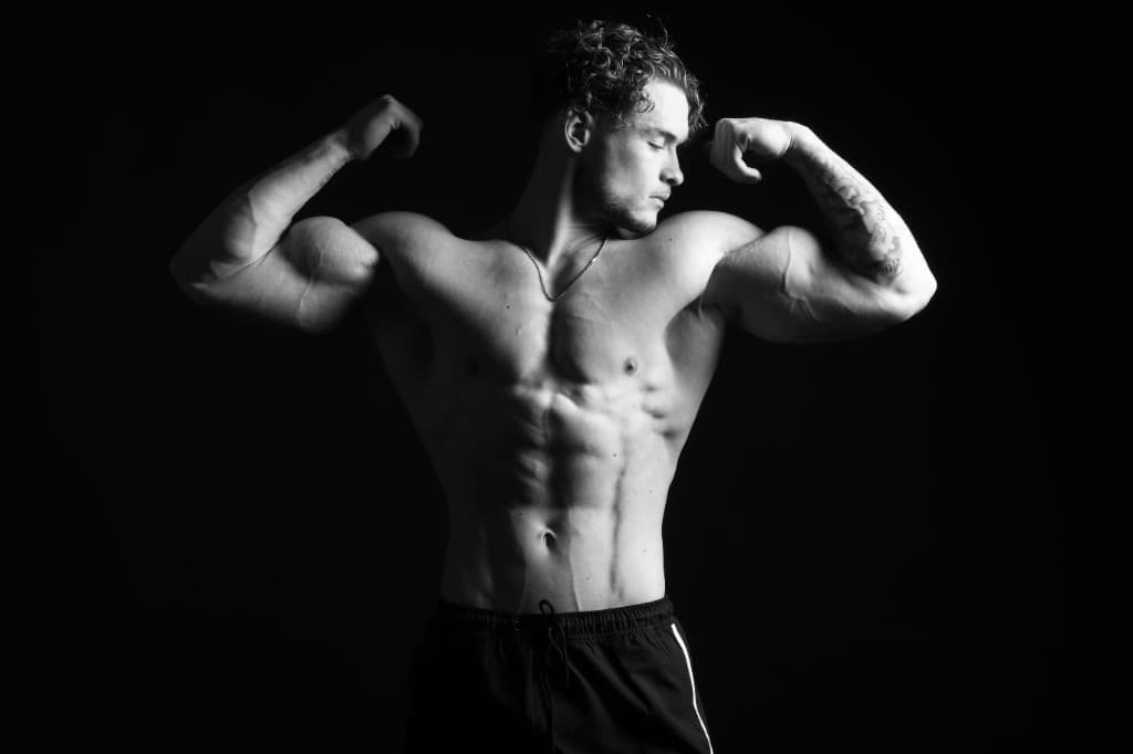 Zwart-wit portret: atletisch figuur, armen gebogen, dramatische belichting op zwarte achtergrond